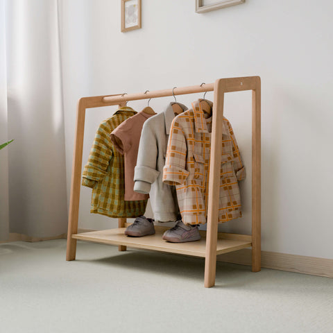 The image displays a Montessori-style clothing rack crafted from natural wood, positioned against a light-colored wall in a brightly lit room. Several children's coats and a pair of shoes are neatly organized on the rack. The coats, hanging from black hangers, vary in patterns and colors, adding a playful and stylish touch to the simple and functional design of the rack. 