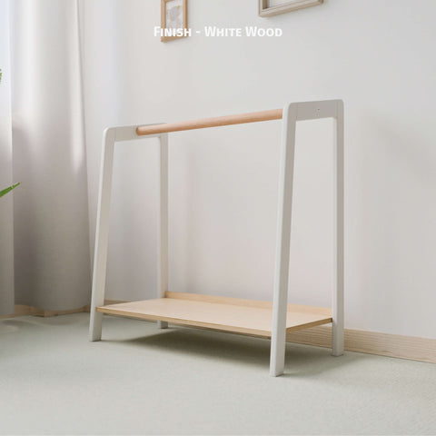 The image displays a Montessori-style clothing rack with a chic white and natural wood finish. The frame is painted white, creating a modern contrast with the natural wood hanging rod and bottom shelf. This minimalist yet functional design fits perfectly in a contemporary nursery or child's room, promoting organization and accessibility for children. Set against a light-colored wall and soft carpeting, the space is warm and inviting. 