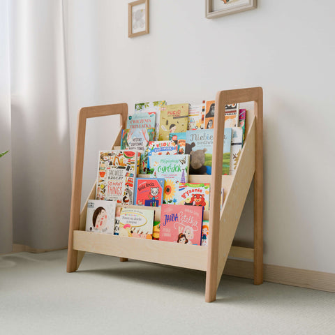 The image features a front-facing Montessori-style bookshelf filled with a vibrant and colorful collection of children's books. The wooden frame of the bookshelf showcases a natural wood finish, enhancing the aesthetic appeal of the room. The shelf is placed against a light-colored wall, softly illuminated by natural light, creating a welcoming and stimulating environment for reading and learning. The design is specifically crafted to display the book covers to encourage young readers' interest.