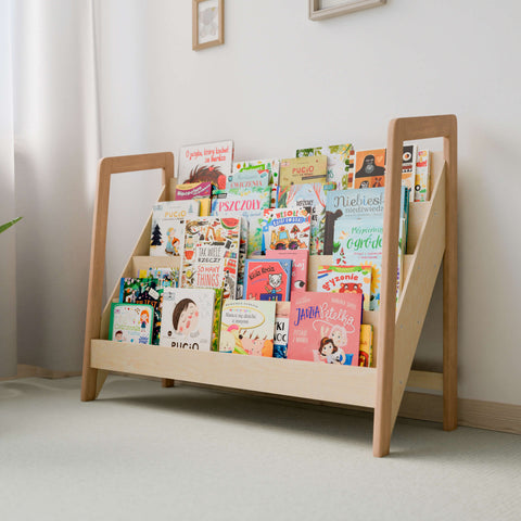 The image shows a large Montessori-style bookshelf made of natural wood. The bookshelf has multiple tiers, displaying a variety of colorful children's books with their covers facing outward for easy selection. The minimalist design fits well in a light-colored, softly lit room, promoting a clean and organized space ideal for a child's play area or bedroom. The natural wood finish enhances the serene and inviting atmosphere, encouraging children to explore and enjoy reading.