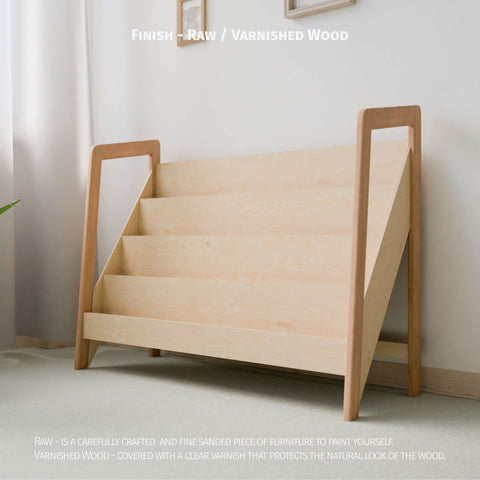 The image shows a large Montessori-style bookshelf made of raw or varnished natural wood. The bookshelf has multiple tiers designed for easy display and access to children's books or toys. The minimalist design fits well in a light-colored, softly lit room, promoting a clean and organized space ideal for a child's play area or bedroom. The natural wood finish enhances the serene and inviting atmosphere, allowing for customization with paint or varnish to protect the wood's natural look.