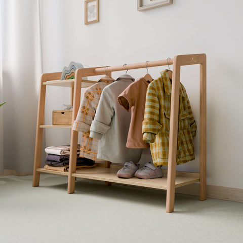 The image shows a large Montessori-style wardrobe made of natural wood. The wardrobe has a hanging rod displaying children's clothes, including jackets and shirts, and two shelves with neatly folded clothes and a pair of shoes. The minimalist design fits well in a light-colored, softly lit room, promoting a clean and organized space ideal for a child's bedroom. 