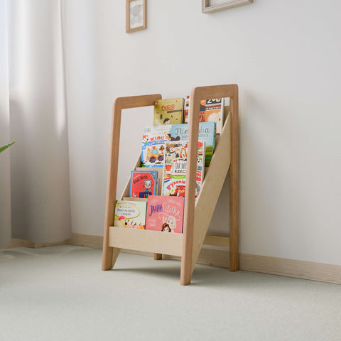 The image depicts a small Montessori-style front-facing bookshelf filled with colorful children's books. The bookshelf is made of natural wood and features a slanted design that displays the covers of the books, making it easy for children to see and select their favorites.