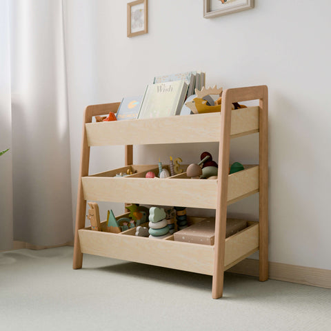  The image depicts a Montessori-style toy organizer made of natural wood, featuring three tiers of open bins. The organizer is filled with various toys and books, promoting easy access and organization for children. The design is minimalist and functional, fitting well in a light-colored, softly lit room. The open bins allow children to see and reach their toys and books easily, encouraging independence and tidy habits. 