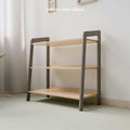The image displays a three-tiered toy storage shelf with a stylish grey and natural wood finish. The structure's grey-painted legs contrast effectively with the light natural wood shelves. Set against a clean, light-colored wall, the setting is serene, lit by natural light filtering through a window to the left, enhancing the colors and giving the space a calm, organized feel. The overlay text specifies the finish as "Grey Wood.