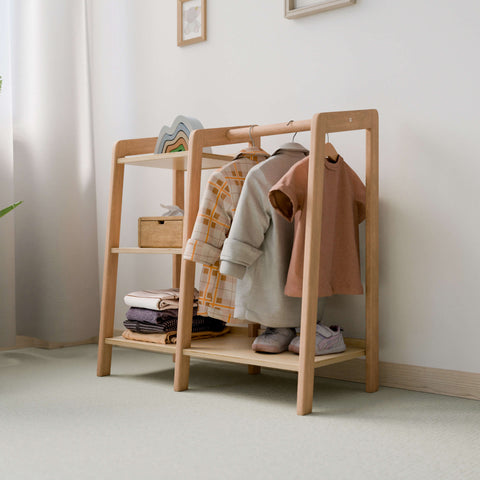 A minimalist children's wardrobe featuring a wooden frame. The wardrobe displays neatly hung clothing, including a checkered shirt, a gray jacket, and a pink dress. The lower shelves hold folded clothes, shoes, and a small wooden box, set in a bright, naturally lit room.