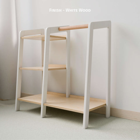 The image showcases an empty Montessori-style wardrobe featuring a white and natural wood finish. The frame is painted white, complementing the natural wood shelves and hanging rod, creating a modern and clean look. This minimalist design includes shelving space for folded clothes and a rod for hanging garments, promoting organization and accessibility for children.