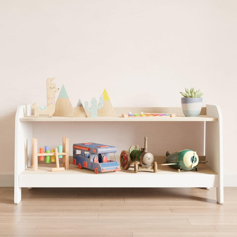 Low and wide montessori toy rack. It has white sides and front and wooden shelves on which children's toys are arranged.