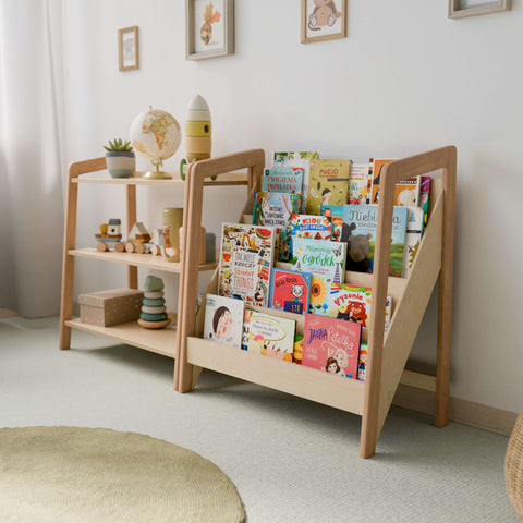 The image shows a Montessori-style setup featuring a bookshelf and a toy storage shelf, both made of natural wood. The bookshelf has multiple tiers displaying a variety of colorful children's books with their covers facing outward for easy selection. The toy storage shelf has three tiers, displaying items such as a globe, a rocket-shaped stacking toy, wooden vehicles, and decorative containers.