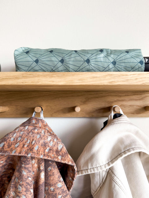 Close-up detail of the oak coat rack showing sturdy wooden hooks holding jackets. The top shelf provides additional storage space, displayed here with a folded umbrella.