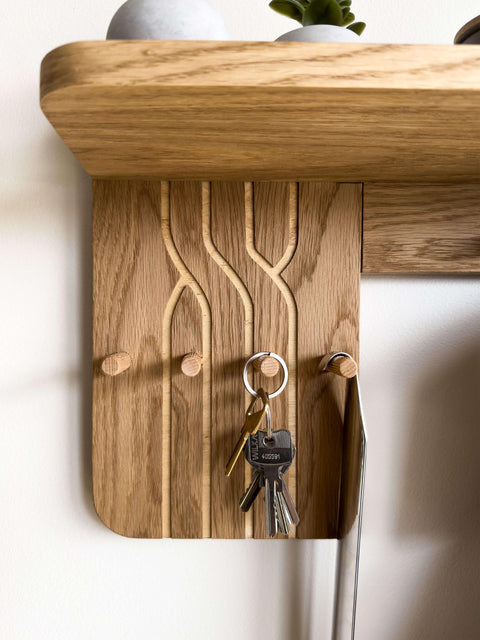 Close-up view of the engraved oak panel on the wall-mounted coat rack. Small wooden hooks serve as a convenient key holder, while the shelf above displays a potted plant.