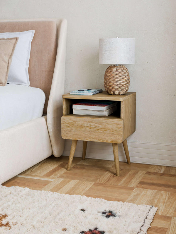 Mid-century oak nightstand with one drawer and open shelf placed beside a bed styled with a woven lamp and books