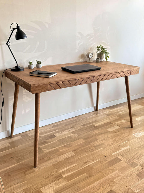 Solid wood desk with geometric carved front details and tapered legs, styled with a laptop and lamp in a modern home office.