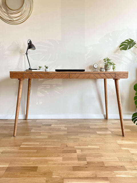 Front view of a handcrafted solid wood desk featuring a geometric carved drawer front and tapered legs in a minimalist workspace.