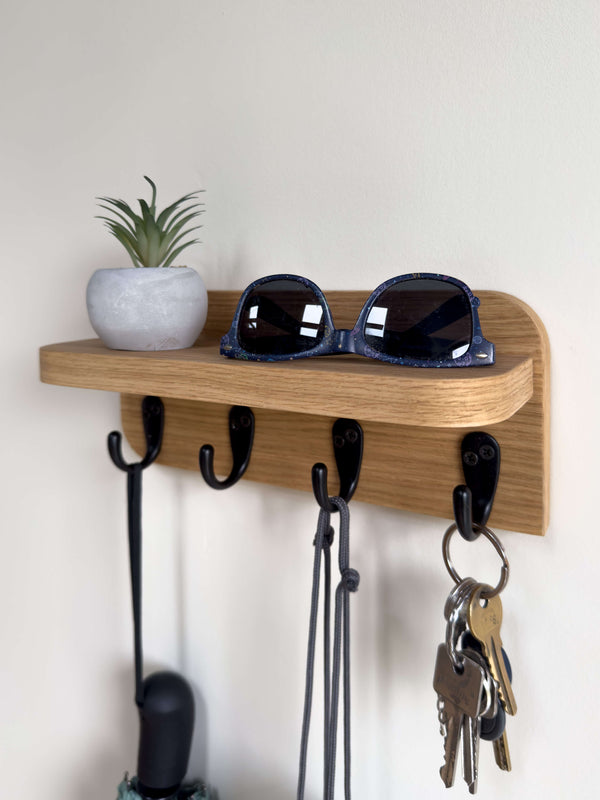 Wall-mounted wooden key holder shelf in natural oak finish with 4 black hooks holding keys and sunglasses