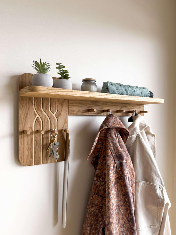 Wall-mounted oak coat rack with shelf installed on a white wall. This entryway organizer features decorative wood engraving, hooks for keys and jackets, and a top shelf for decor.
